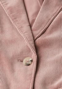 Sametový saka v jemně růžové barvě s širokými klopami, které má velký krémový knoflík a jemný texturovaný vzor. Podšité hladkým materiálem.