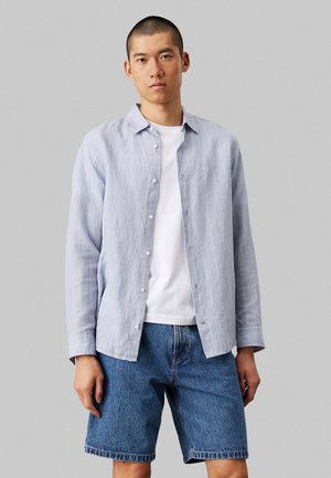 Camicia azzurra chiara a maniche lunghe con tessuto strutturato, abbinata a pantaloni corti di denim. La camicia è indossata sopra una maglietta bianca.