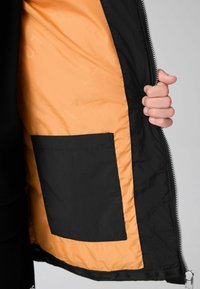 Černá bunda s oranžovým podšívkou, obsahující velkou vnitřní kapsu. Hladká textura, zipové zapínání a těsný střih u dolního okraje.