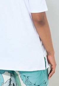 Camiseta de algodón blanca con aberturas laterales, con mangas cortas y un corte relajado. Se ve una mano que muestra un tatuaje. Textura suave.