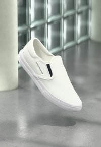 Λευκά παντόφλες από καμβά με λεία υφή, ελαστικές πλαϊνές λεπτομέρειες και διακριτικό μαύρο λογότυπο στην εξωτερική πλευρά. Λαστιχένια σόλα.