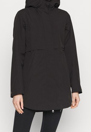 Femme portant une veste imperméable noire à capuche avec manches longues et fermeture éclair frontale, associée à un legging noir.