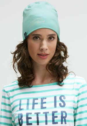 Woman with curly brown hair wearing a turquoise beanie and a white shirt with turquoise stripes and "LIFE IS BETTER" text.