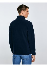 Mężczyzna stojący bokiem, ubrany w granatową polarową kurtkę z wysokim kołnierzem i niebieskie dżinsy na jednolitym jasnym tle.