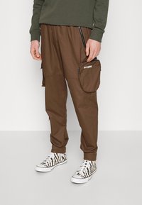 Bruine cargobroek met zijritsen en een grote zak. Gecombineerd met zwart-wit gestreepte sneakers met een zebra-patroon. Zachte stoftextuur.