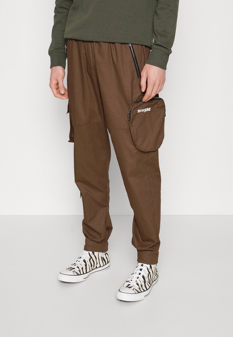 Bruine cargobroek met zijritsen en een grote zak. Gecombineerd met zwart-wit gestreepte sneakers met een zebra-patroon. Zachte stoftextuur.