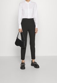 Bílá dlouhá košile s límcem, černé kalhoty s vysokým pasem, černá kabelka a masivní černé boty se stříbrnými kovovými detaily.