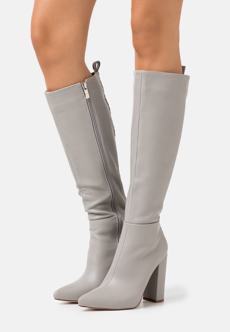 Botas de cuero gris claro hasta la rodilla con cremallera lateral, tacones de bloque y punta afilada, usadas sobre piernas desnudas.