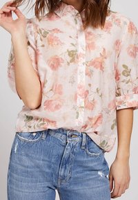 Lichtroze bloemenknopshirt met opgerolde mouwen. Zachte, sheer stof met pastelrozen en groene bladeren, in combinatie met blauwe versleten jeans.