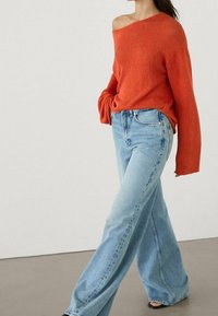 Pull tricoté orange vif à épaules dénudées avec manches longues, associé à un jean bleu clair à jambes larges. Textures lisses, coupe décontractée.
