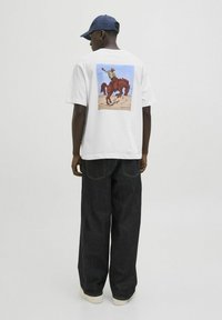 Camiseta blanca con un gráfico colorido de un vaquero montando un caballo en la parte de atrás, combinada con pantalones de mezclilla oscuros y una gorra azul.