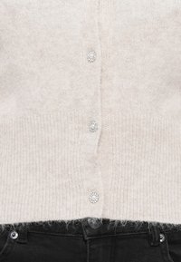 Cardigan en maille beige clair avec boutons en perles et strass, associé à un jean en denim noir, présenté de la taille à la moitié du torse.