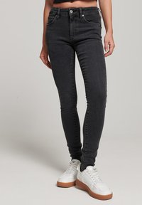 Jeans ajustados de cintura alta en negro, fabricados en denim, con cinco bolsillos, cremallera y detalles de costura sutiles, combinados con zapatillas blancas.