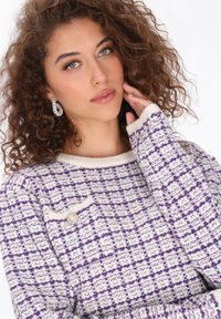 Kötött pulóver, fehér és lila kockás mintával, kerek nyakkivágással és díszes brosstűvel a mellrészen. Puha tapintású.