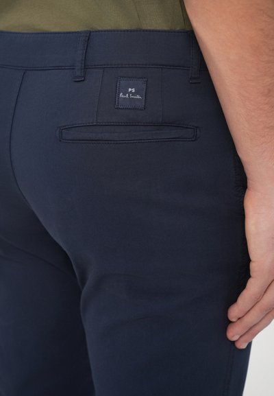 Tmavomodré nohavice s hladkou textúrou, s zadným vreckom, na ktorom je obdĺžniková etiketa s nápisom "PS Paul Smith".