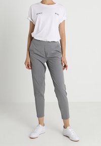 Persoon in een wit T-shirt met de tekst "WACHT NIET", grijze slim-fit broek en witte sneakers, staand voor een effen witte achtergrond.