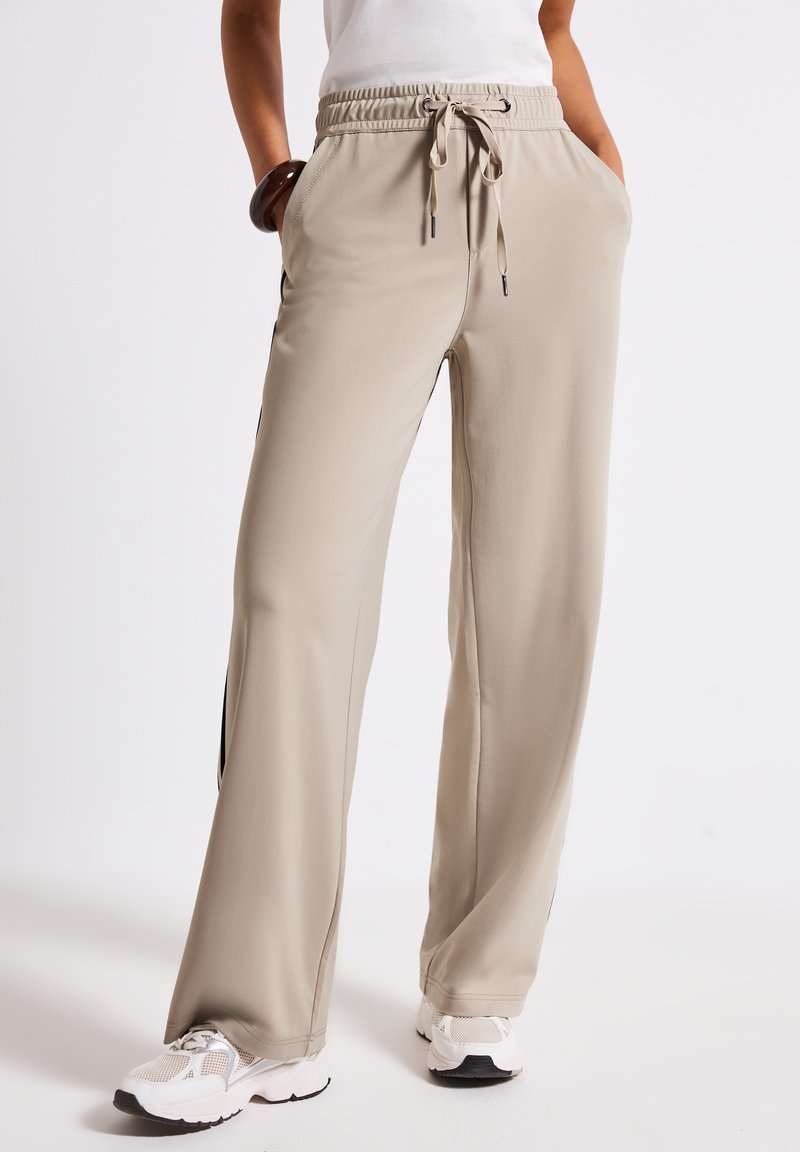 Beige, loose-fit sweatpants with an elastic waistband and drawstring, featuring side pockets and black side stripes. Smooth texture.