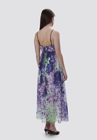 Lungo abito floreale in tessuto trasparente, con motivi viola e verdi, spalline nere regolabili e una silhouette fluida. Tacchi neri.