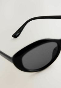 Lunettes de soleil noires en forme ovale, dotées de verres sombres et de montures en plastique. La branche présente un texte en relief indiquant la certification CE et l'origine.