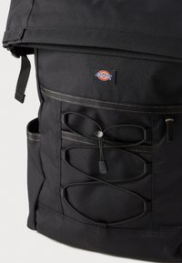 Mochila preta feita de tecido durável com design de topo enrolável, apresentando um patch com logo, detalhes em cordão elástico e bolsos laterais exteriores.