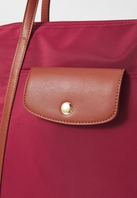 Maroon nylon tote bag with brown leather flap and gold snap closure. Features smooth texture and contrast stitching along the edges.