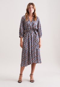 Gemustertes Midikleid aus blauer, roter und weißer Stoff. Hat einen V-Ausschnitt, Ballonärmel, eine taillierte Taille mit einem Gürtel und seitliche Taschen.