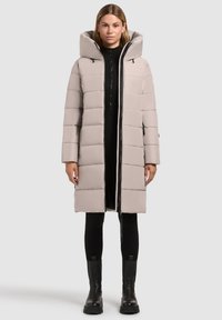 Polstret beige frakke med høj krave og stor hætte, der har en fuld frontlynlås og lynlåslommer, båret over et sort outfit.