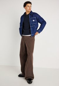 Donkerblauwe denim jack met een voorzak, gelaagd over een donkere trui. Bruine wijde broek. Zwarte schoenen maken de outfit compleet.