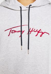 Sweat à capuche gris clair avec cordons noirs et texte en cursive rouge à sequins sur la poitrine.