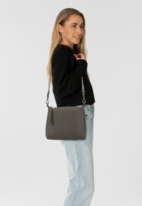 Bolso de hombro cruzado de piel gris con una textura suave, que cuenta con una larga correa ajustable, detalles de cremallera y un diseño minimalista.