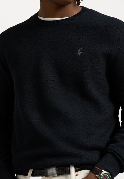 Pull en tricot noir avec un col rond et des poignets côtelés, arborant un petit logo brodé sur la poitrine. Porté par-dessus un pantalon de couleur claire.