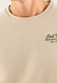 T-shirt in cotone beige con scollatura rotonda e maniche corte. Presenta sulla parte anteriore una scritta verde che recita "Club Miami Beach (555) 125".