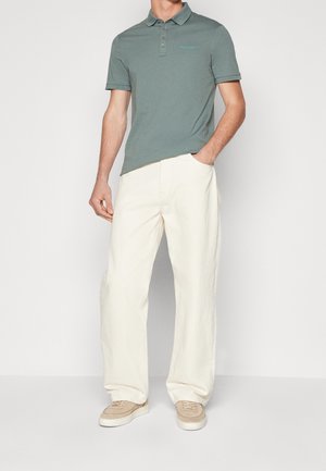 Homme portant un polo vert à manches courtes, un pantalon large crème et des baskets beige, debout avec une main dans la poche, sur fond blanc.