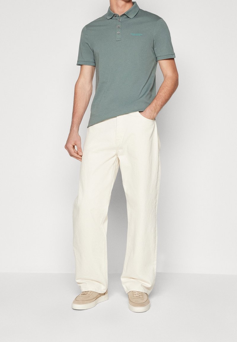 Homme portant un polo vert à manches courtes, un pantalon large crème et des baskets beige, debout avec une main dans la poche, sur fond blanc.