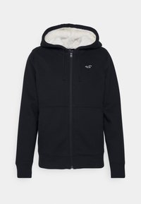 Svart zip-up huvtröja med ett foder av konstpäls i huvan, med en ficka på framsidan och en liten broderad logotyp på bröstet. Slät tygstruktur.