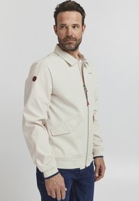 Light cream jacket with a zip front, pointed collar, side pockets, elastic cuffs, and embroidered patch on the sleeve. Pair of dark jeans.