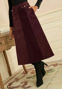 Gonna midi in corduroy bordeaux con un frontale piatto, due tasche laterali e due bottoni metallici decorativi, abbinata a stivaletti neri a punta.