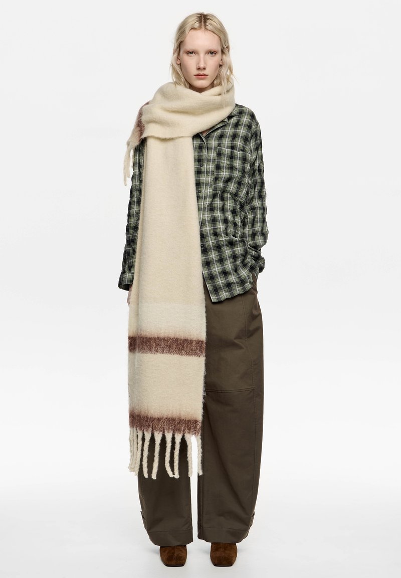 Beżowy wełniany szalik z brązowymi paskami, z frędzlami na końcach, noszony na zielonej, kraciastej koszuli i luźnych brązowych spodniach. Miękka tekstura i ciepły fason.