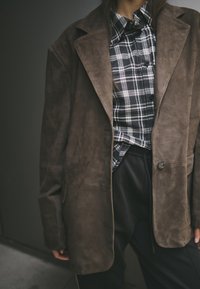 Bruine suedé oversized blazer met gegoten kleppen, gedragen over een zwart-wit geblokt shirt, met knopen en een relaxte pasvorm.