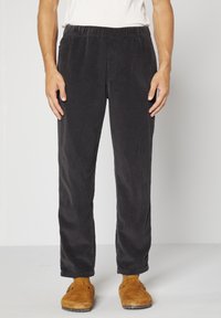 Pantalon en velours côtelé noir avec des côtes verticales, taille élastique, jambes fuselées et poches latérales. Associé à des sabots en daim marron.