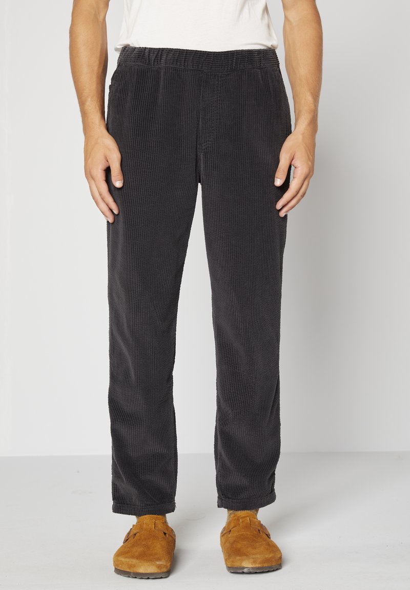 Pantalon en velours côtelé noir avec des côtes verticales, taille élastique, jambes fuselées et poches latérales. Associé à des sabots en daim marron.