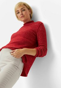 Rød, tekstureret turtleneck sweater med lange ærmer, vist over beige bukser, har en afslappet pasform og sideslids for ekstra komfort.
