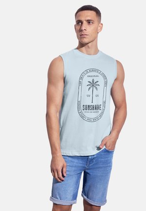Hellblaues ärmelloses Shirt mit einem Grafikdesign einer Palme und Text. Kombiniert mit hochgekrempelten Jeansshorts. Lässiger und entspannter Schnitt.