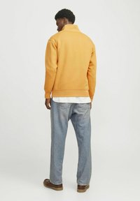 Felpa arancione con colletto alto, abbinata a jeans di denim blu chiaro. Il tessuto appare morbido, con una vestibilità rilassata e polsini a coste.