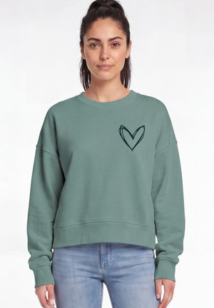 Jonge vrouw met donker haar in een paardenstaart, die een groene trui met een zwart hart op de borst en lichtblauwe spijkerbroek draagt.