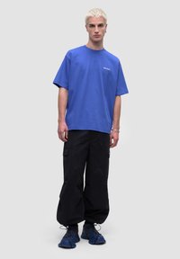 Niebieska koszulka z krókim rękawem, z okrągłym dekoltem i detalem z logo, w połączeniu z luźnymi czarnymi spodniami cargo i niebieskimi butami sportowymi.