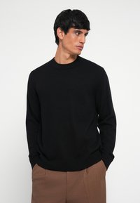 Black knit sweater with a crew neck and long sleeves, featuring a smooth texture and a relaxed fit. Paired with brown trousers.