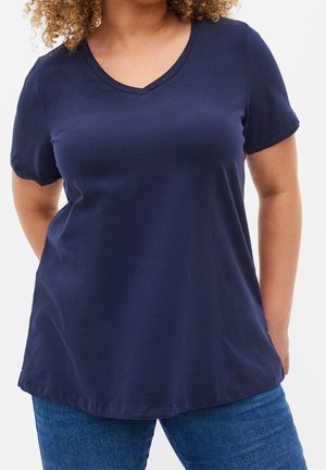 Woman wearing a navy blue V-neck short-sleeve T-shirt and blue jeans, standing with hands behind her back.