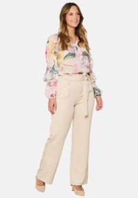 Blusa floreale con colori pastello, maniche lunghe e dettagli a volant, abbinata a pantaloni beige a vita alta con cintura da annodare.
