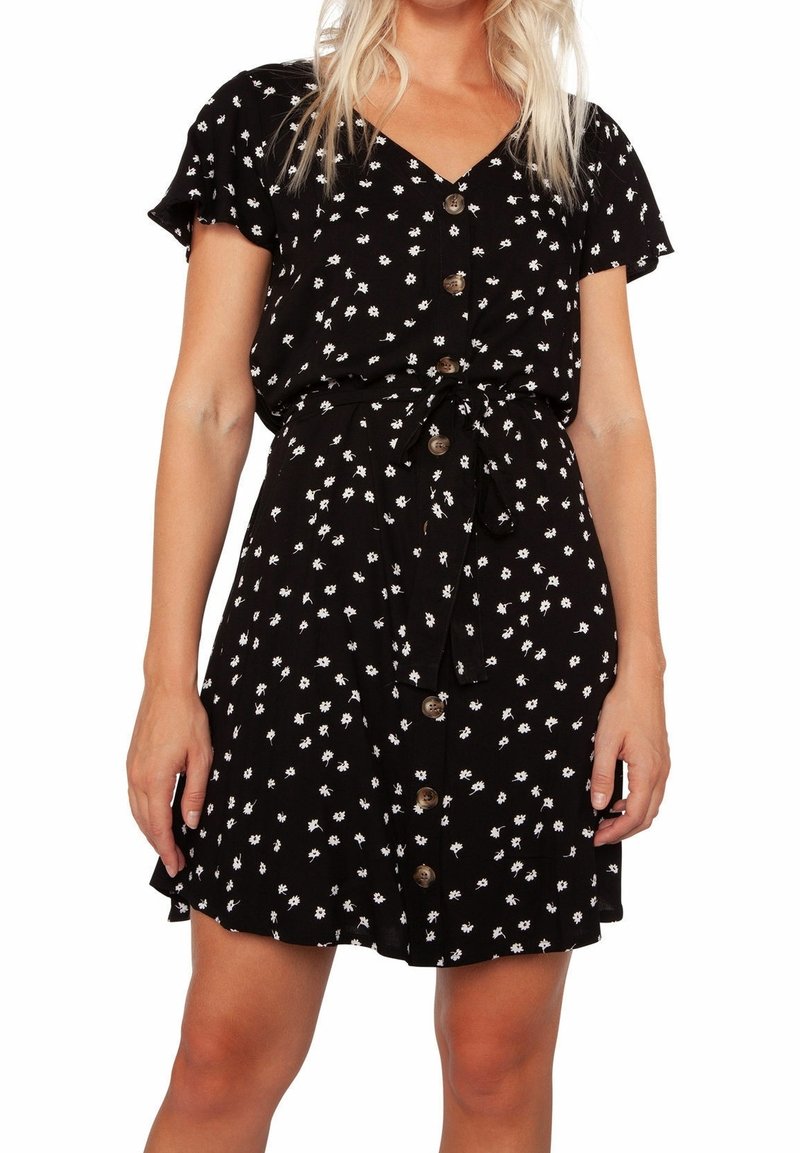 Femme portant une robe noire à manches courtes avec un imprimé floral blanc, des boutons à l'avant et une ceinture nouée à la taille, debout devant un fond blanc.